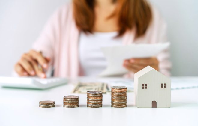 Mujer calculando el impuesto de inmueble con monedas y modelo de casa